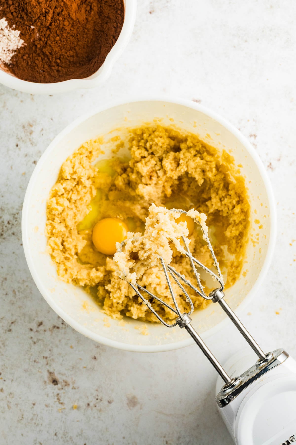 A mixing bowl with creamed butter and sugar, an egg added on top, a hand mixer resting inside, and a bowl of cocoa powder in the background.