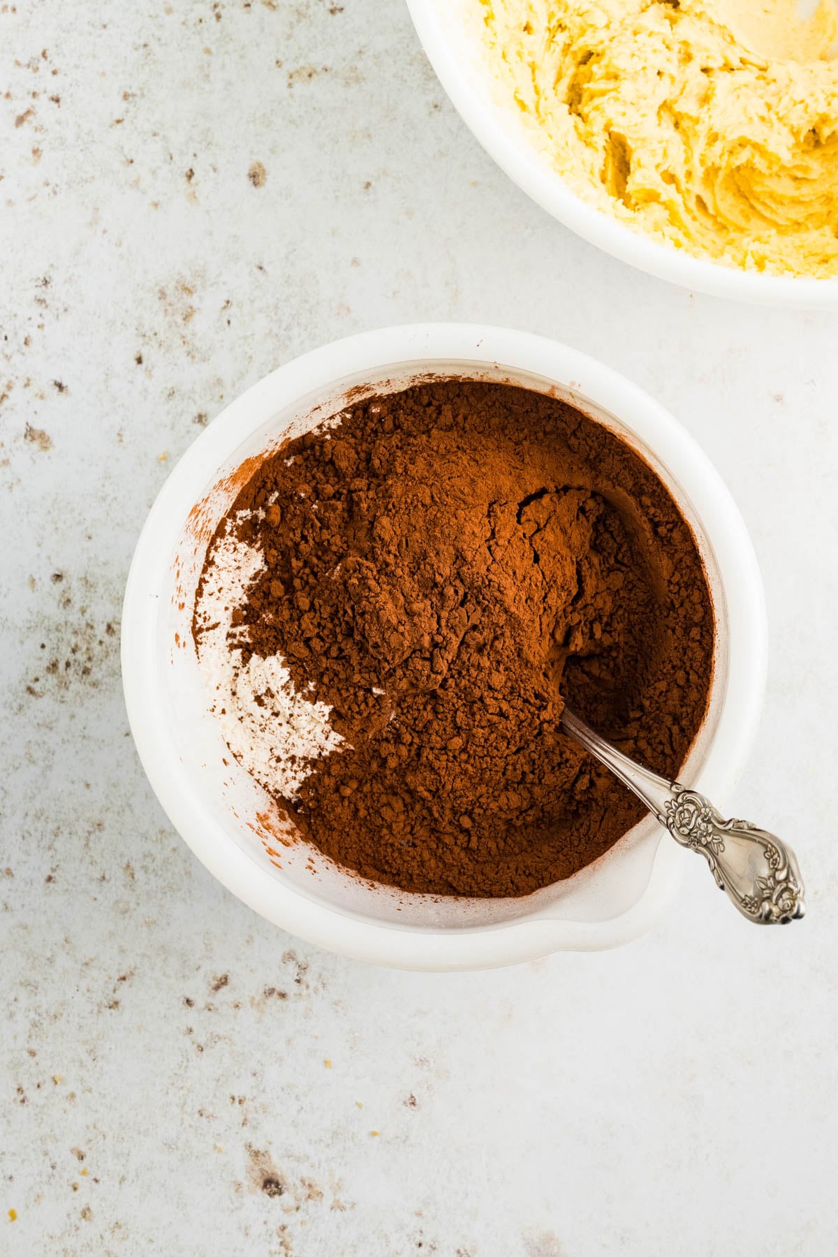 A white bowl filled with cocoa powder and a metal spoon next to a bowl of yellow batter on a light surface.
