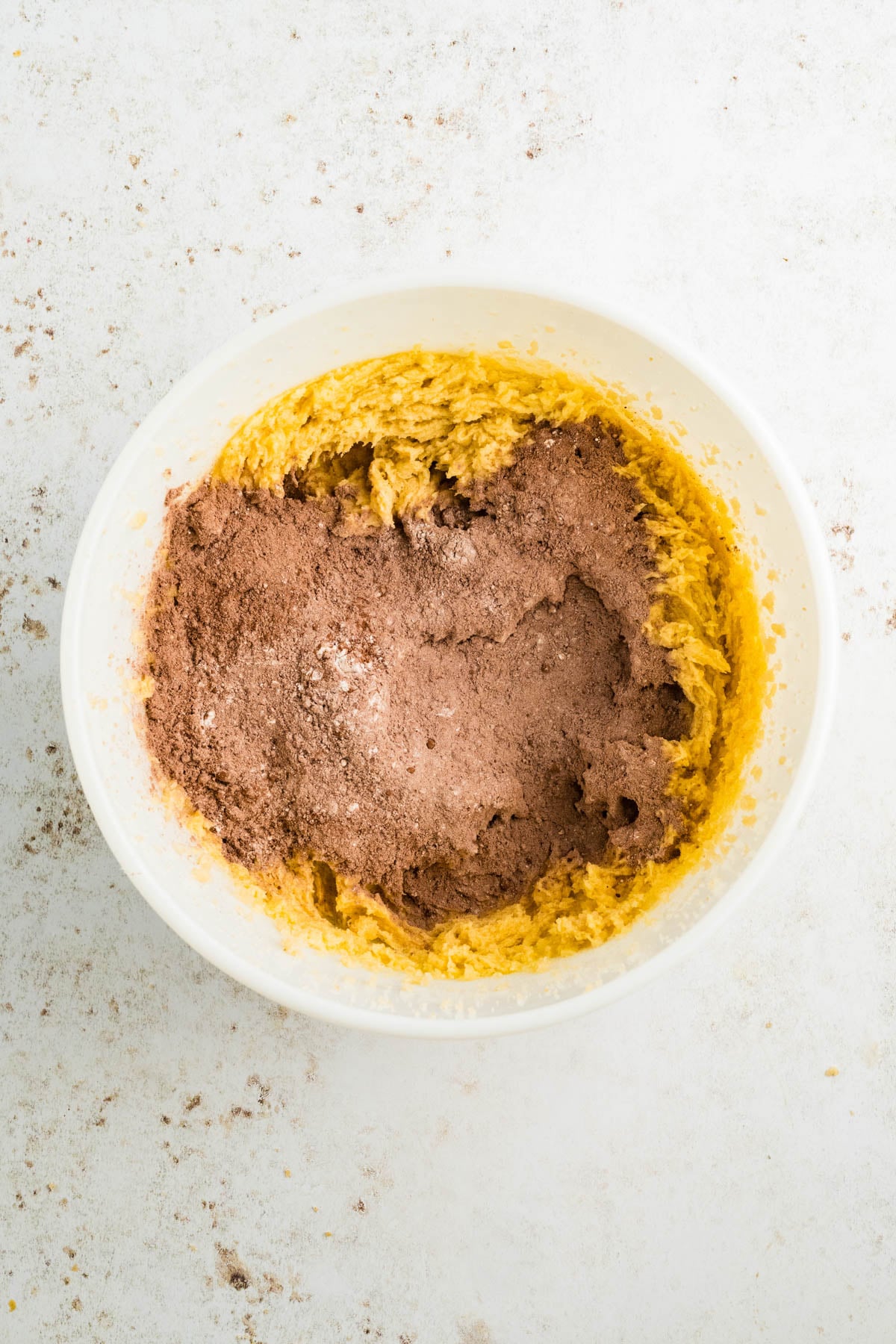 A white bowl containing partially mixed yellow batter and cocoa powder sitting on a light surface.