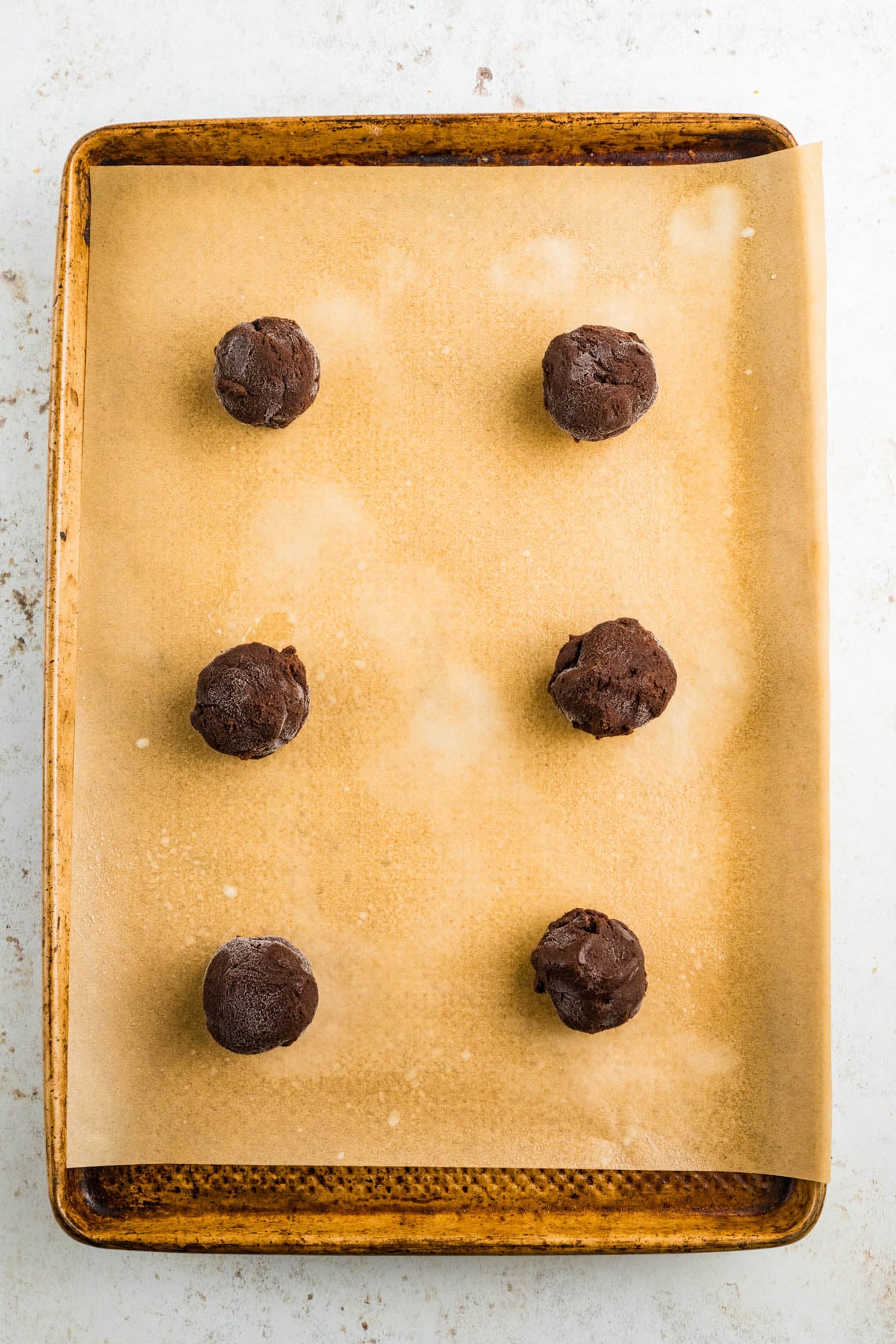 Six portions of chocolate cookie dough are arranged on a parchment-lined baking sheet, ready to be baked.