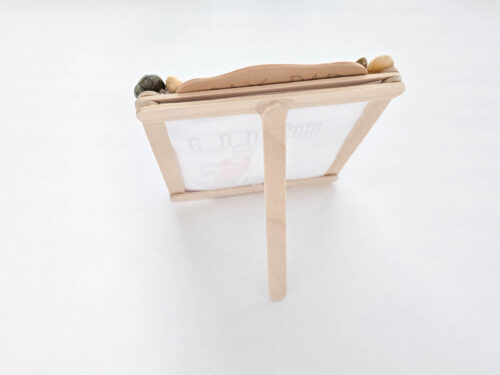 A small picture frame made of popsicle sticks and decorated with pebbles is standing upright on a white surface.