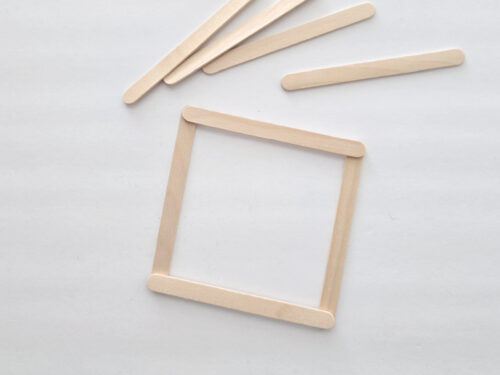Four wooden craft sticks forming a square on a white surface, with four additional sticks lying above the square.