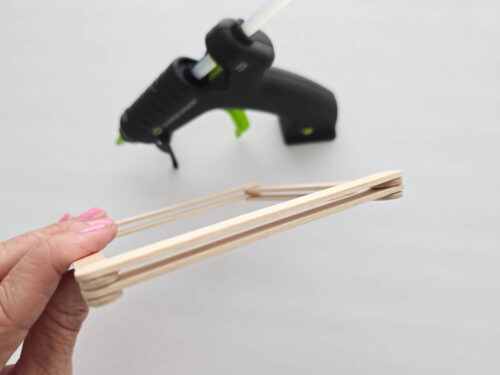 A hand holds a small, rectangular frame made of popsicle sticks; a black hot glue gun is visible in the background on a white surface.