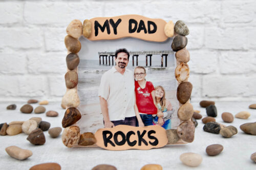 A framed photo decorated with stones shows a man, a woman, and a young girl on a beach. The frame reads "MY DAD ROCKS." Loose stones are scattered around the frame.