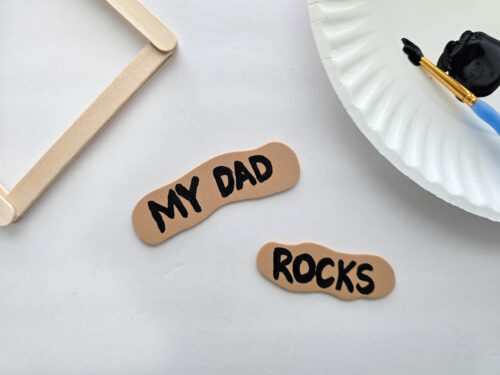 Two tan foam cutouts with black text read "MY DAD" and "ROCKS" on a white surface, next to craft sticks, a paintbrush, a paper plate, and black paint.