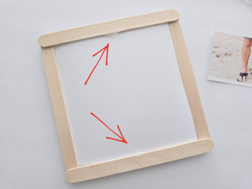 A square frame made of wooden popsicle sticks with a blank white insert, featuring two red arrows pointing to glue spots at the top and bottom.