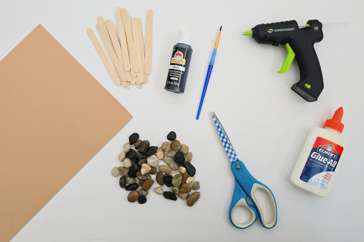 Brown craft foam, wooden sticks, black paint, paintbrush, hot glue gun, small rocks, blue scissors, and white glue arranged on a white surface.