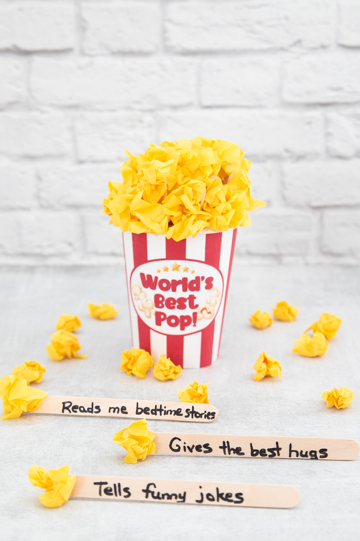 A striped popcorn container labeled "World's Best Pop!" filled with yellow paper, with popsicle sticks reading "Reads me bedtime stories," "Gives the best hugs," and "Tells funny jokes.