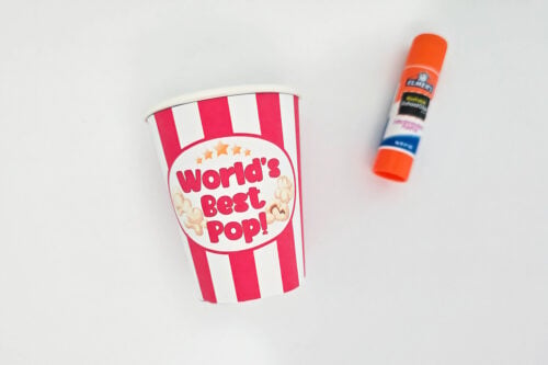 A striped paper cup labeled "World's Best Pop!" sits next to an orange Elmer's glue stick on a white surface.