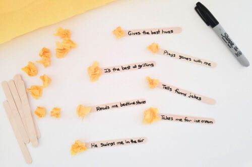 Several craft sticks with handwritten messages such as "Gives the best hugs" and "Takes me for ice cream" are arranged on a table with a marker and crumpled yellow paper.
