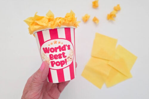 A hand holds a red and white striped cup labeled "World's Best Pop!" filled with crumpled yellow tissue paper; extra tissue paper pieces are on a white surface.