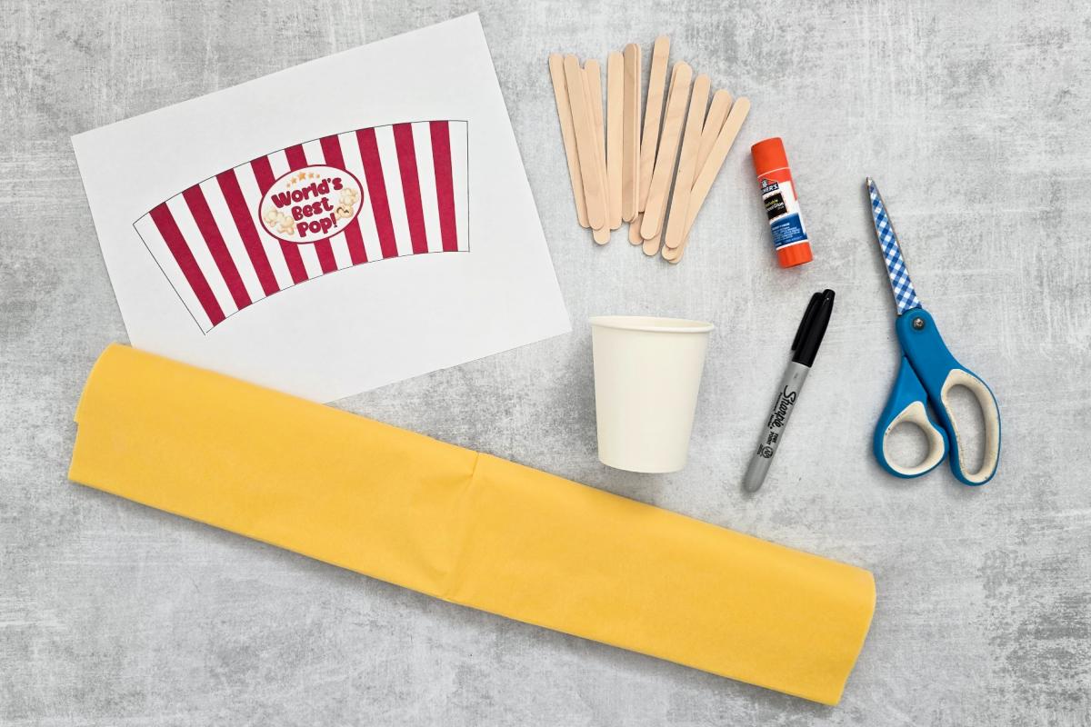 Materials for a craft project: printed paper label, wooden sticks, glue stick, white paper cup, black marker, yellow tissue paper, and blue-handled scissors on a gray surface.