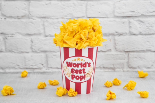 A striped popcorn container labeled "World's Best Pop!" is filled with and surrounded by crumpled yellow paper resembling popcorn.