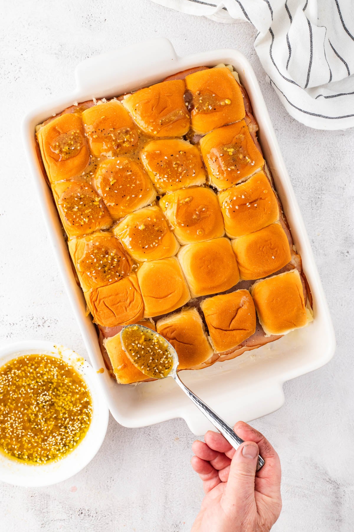 A hand spreads a mustard and seed sauce over baked sliders in a white baking dish, with a bowl of sauce nearby on a white surface.