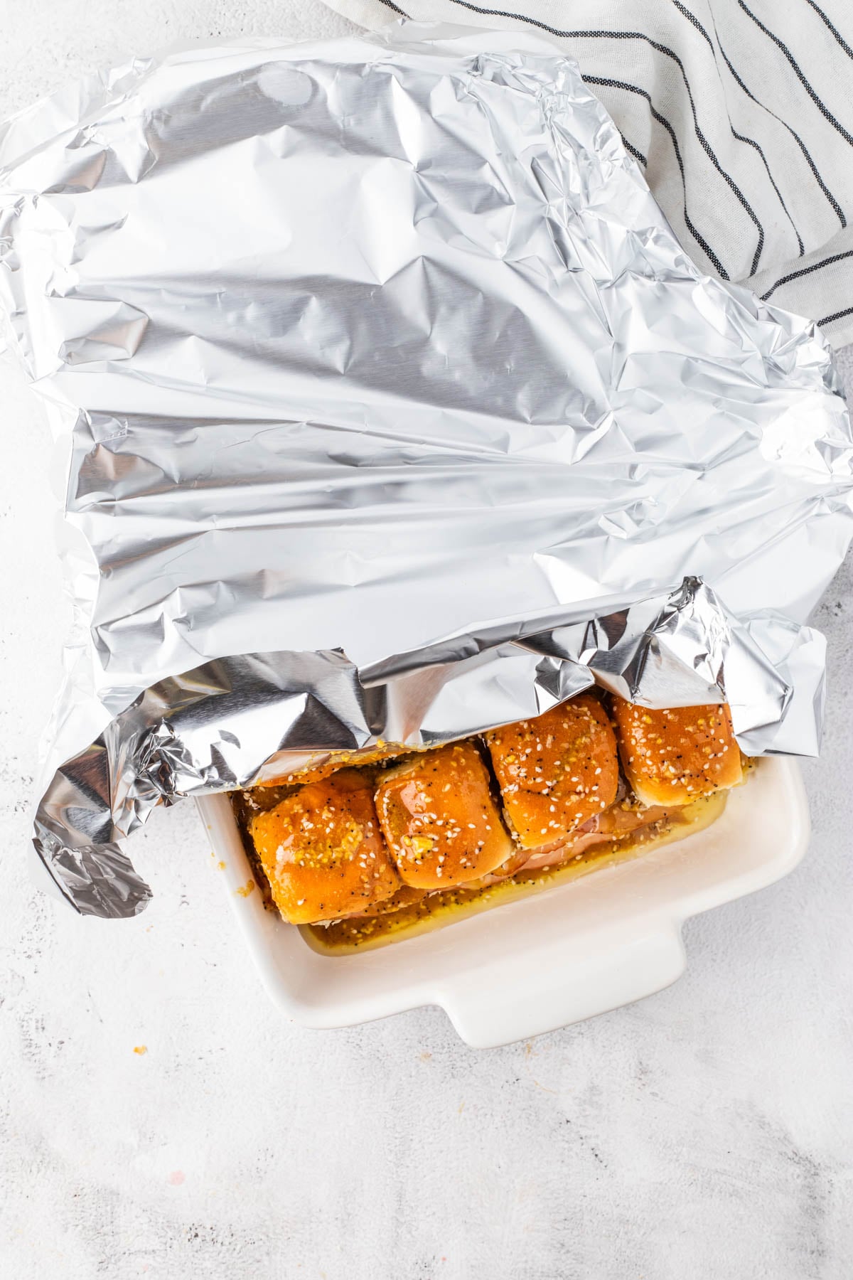 A white baking dish contains six slider sandwiches topped with sesame seeds, partially covered with a sheet of aluminum foil. A striped kitchen towel is nearby.