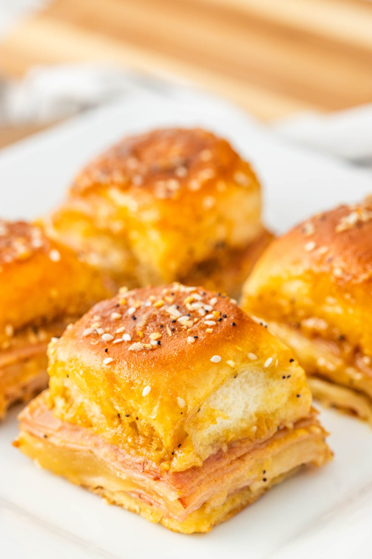Close-up of four baked ham and cheese sliders on a white plate, topped with sesame seeds and seasoning.