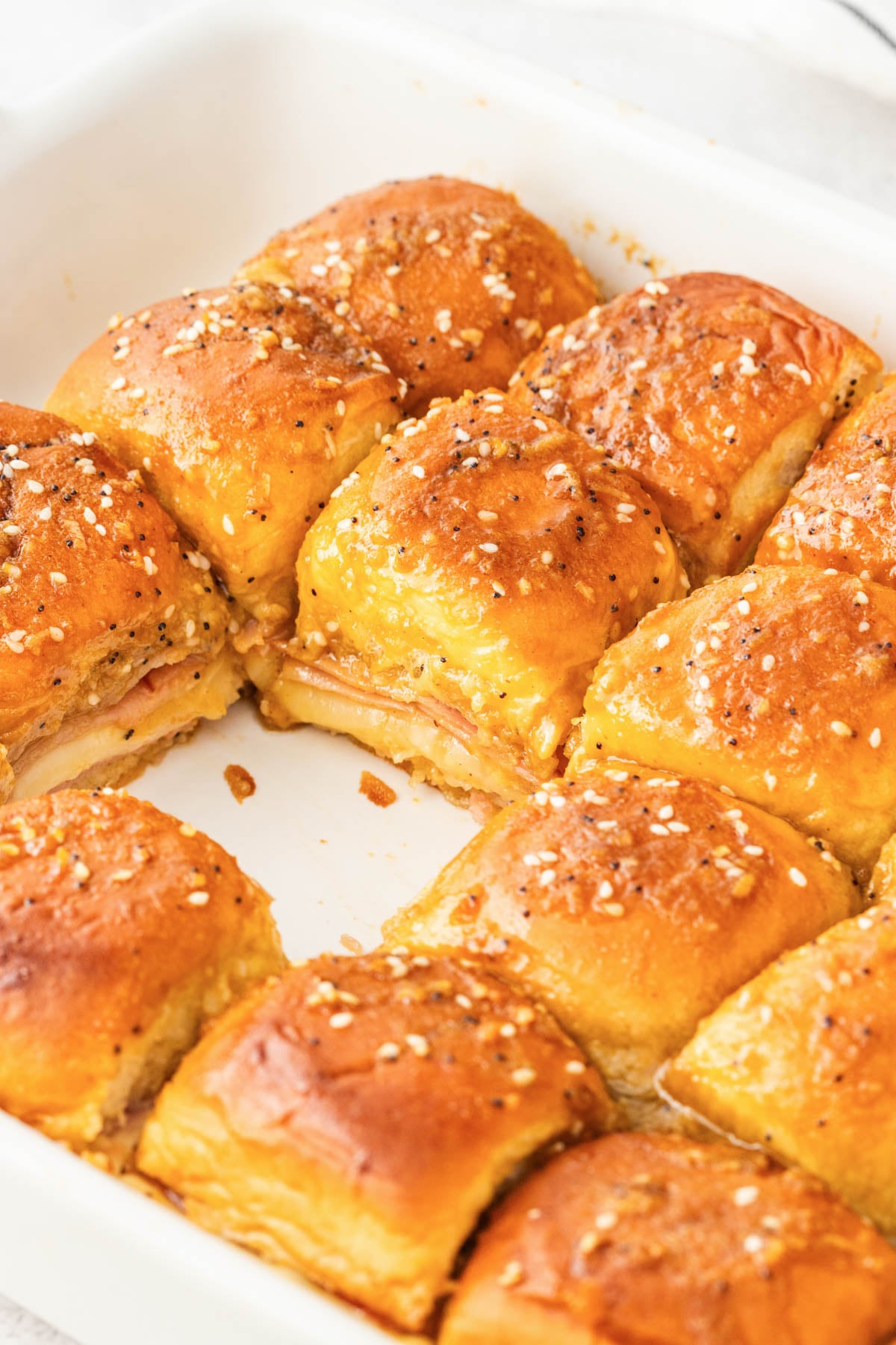 A white baking dish filled with golden brown ham and cheese sliders topped with melted butter and seasoning.