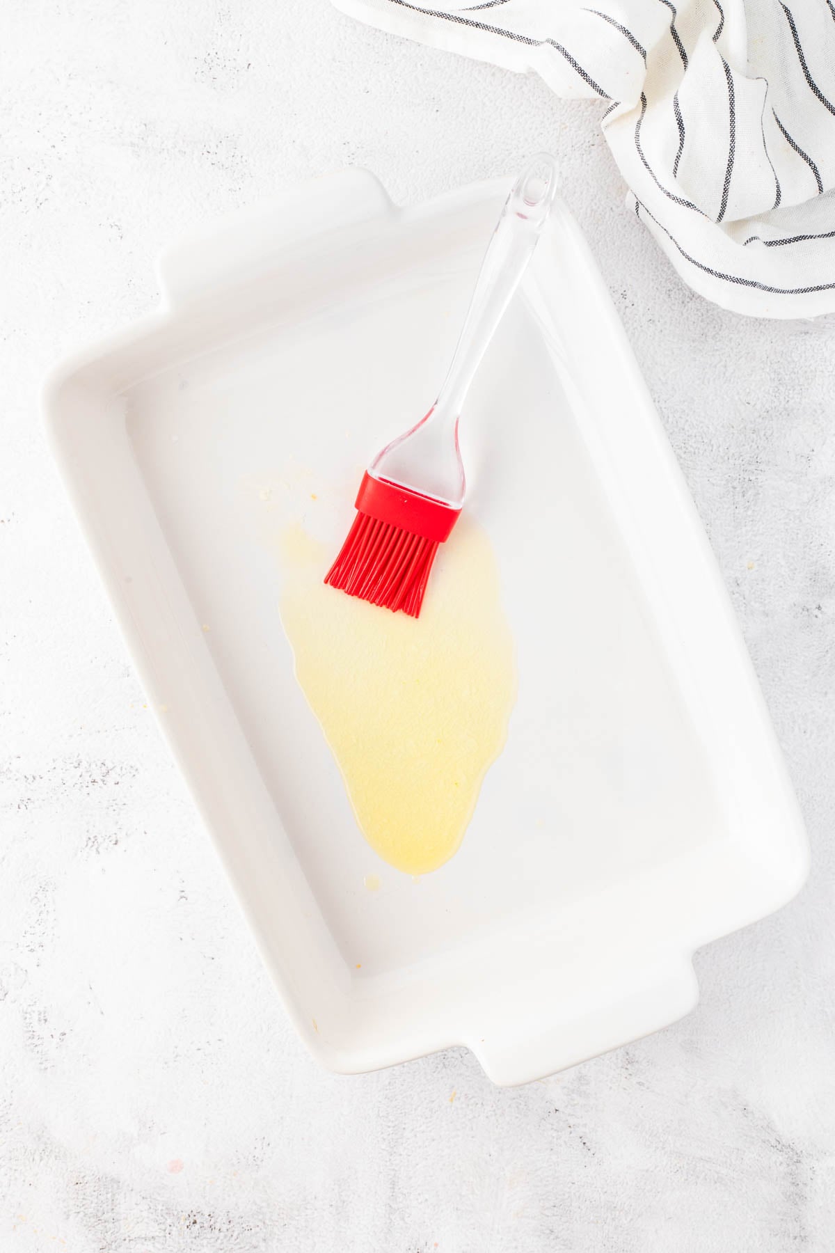 A red silicone pastry brush rests in a white rectangular baking dish with a layer of oil spread across the bottom. A striped cloth is partially visible in the corner.