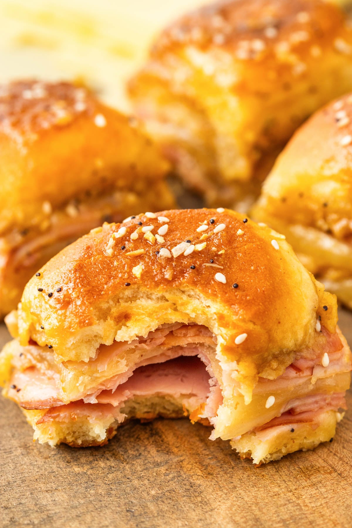 A ham and cheese slider with a sesame seed bun has a bite taken out, showing layers of ham and melted cheese. Other sliders are in the background on a wooden surface.