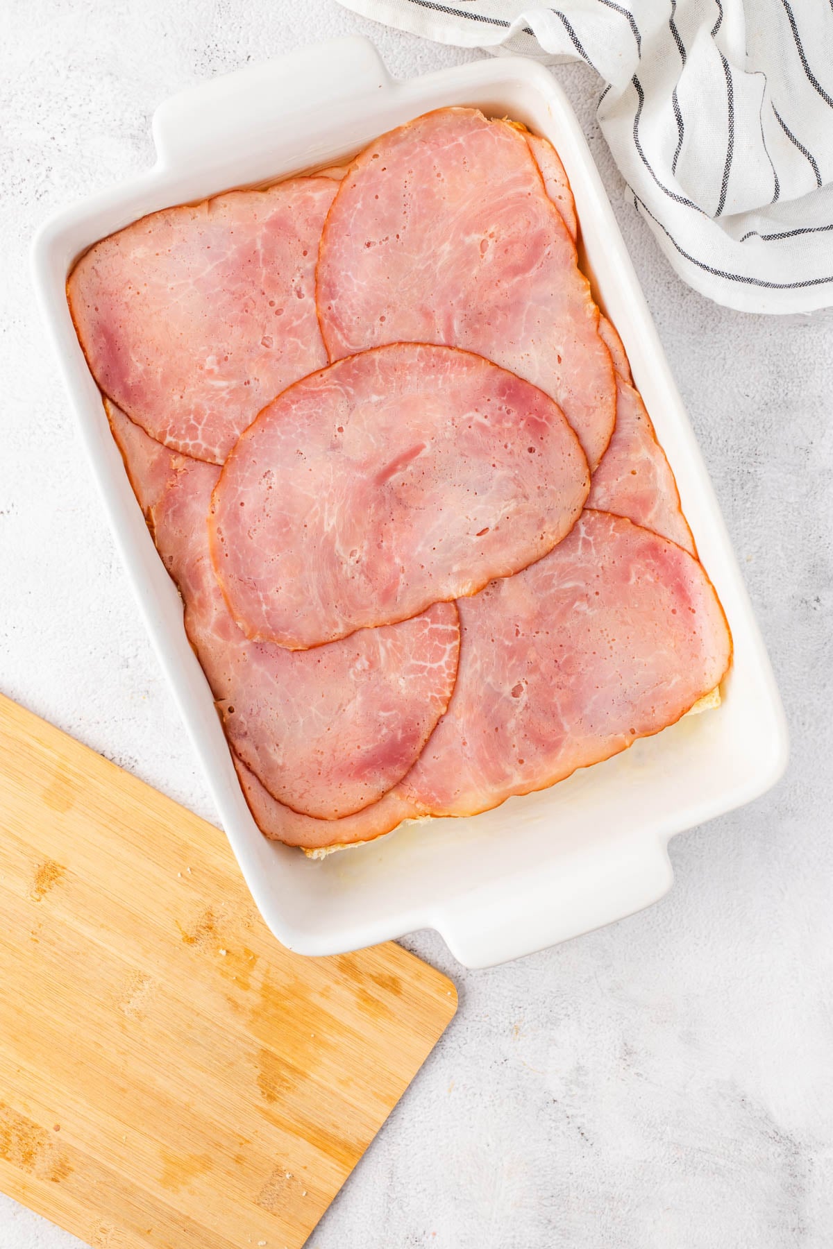 Rectangular white baking dish with overlapping slices of ham laid flat inside, next to a striped towel and a wooden cutting board on a light surface.
