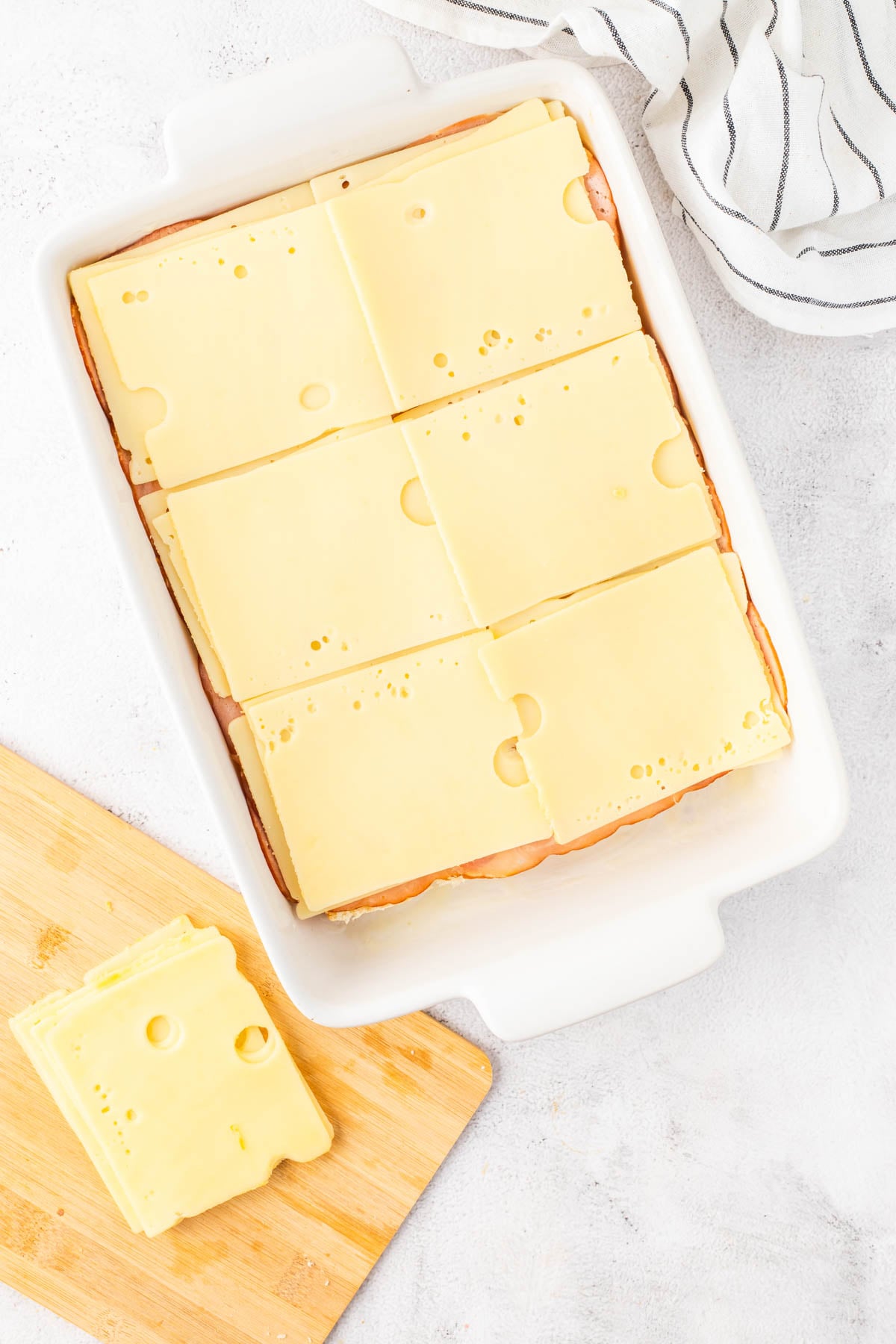 A white baking dish filled with layers of deli ham and Swiss cheese slices, with extra cheese on a wooden cutting board nearby. A striped towel is in the upper right corner.