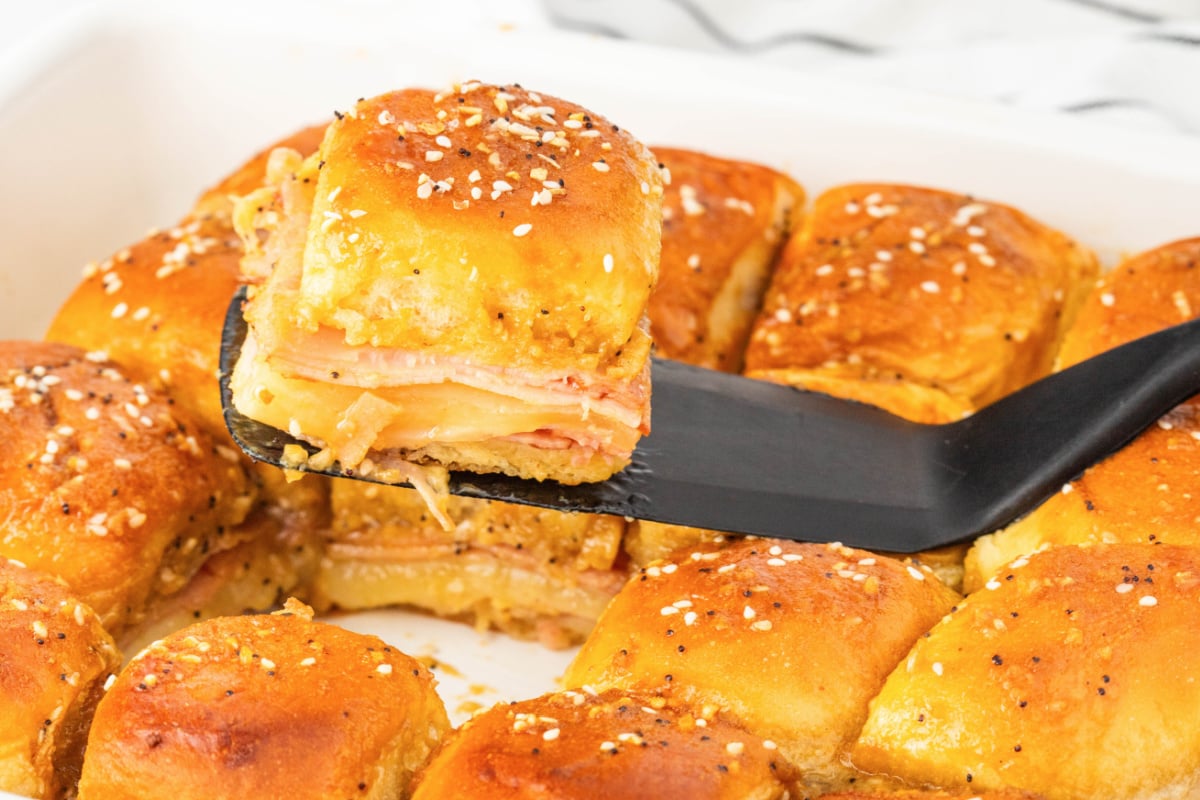 A black spatula lifts a ham and cheese slider with golden, seasoned buns from a baking dish filled with similar sliders.
