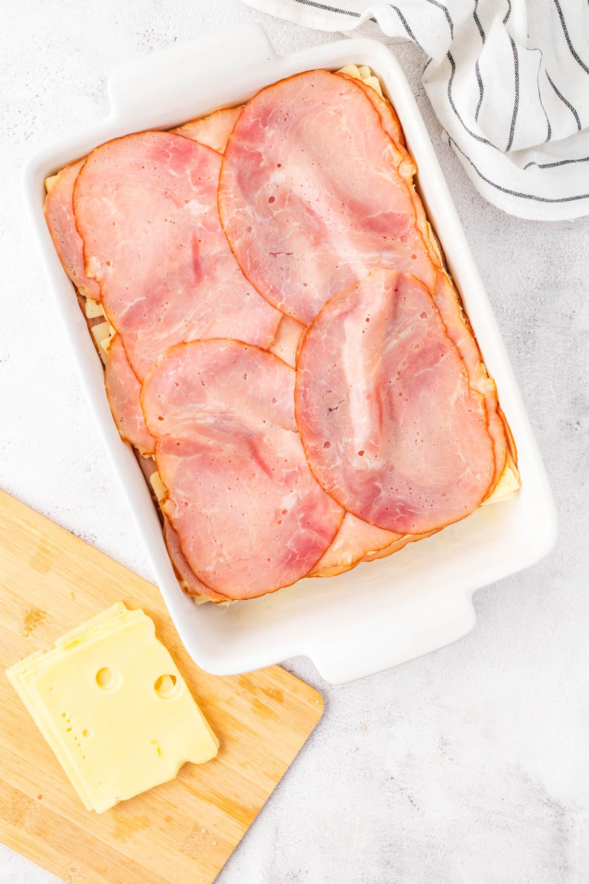 A white baking dish with layers of ham over a casserole, next to a wooden board with slices of Swiss cheese and a striped cloth.