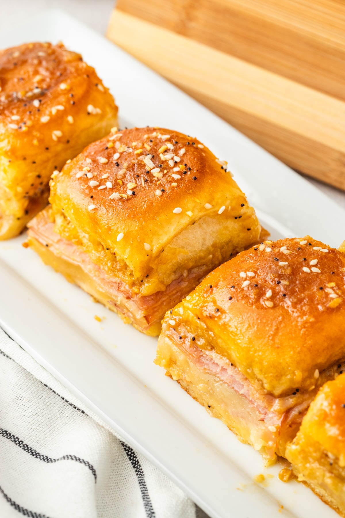 Three ham and cheese slider sandwiches with sesame seed topping are arranged on a white rectangular plate.