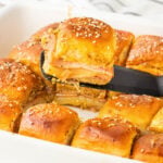 A spatula lifts a ham and cheese slider with a golden, seasoned bun from a baking dish filled with similar sliders.