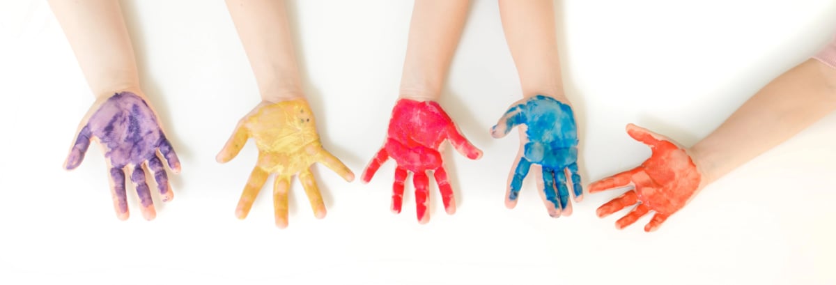 Five children's hands are spread out on a white surface, each palm covered in different colored paint: purple, yellow, red, blue, and orange.