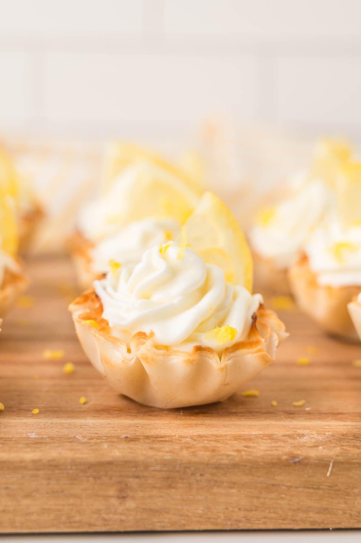 Mini phyllo tart filled with lemon cheesecake and topped with a swirl of whipped topping and a slice of lemon, placed on a wooden surface.