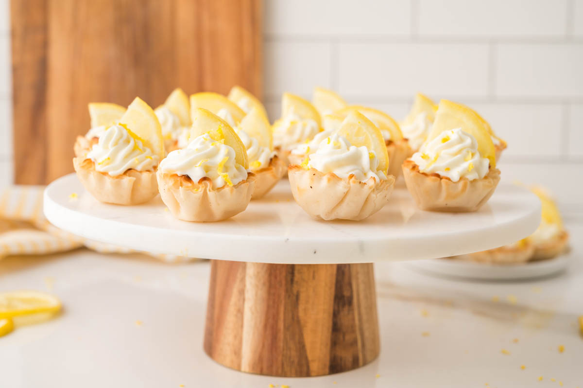 Mini lemon cheesecakes and lemon slices are arranged on a round marble and wood cake stand.