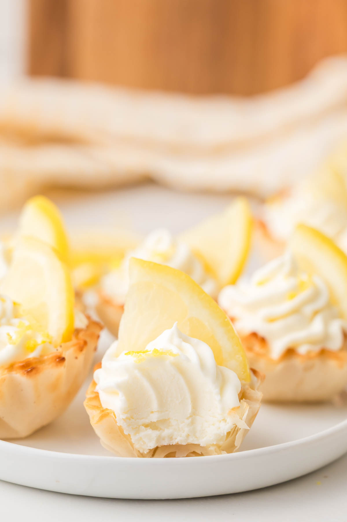 Mini lemon cheesecake bites with whipped cream and lemon slices on top, arranged on a white plate. One mini cheesecake in the front has a bite taken out of it.