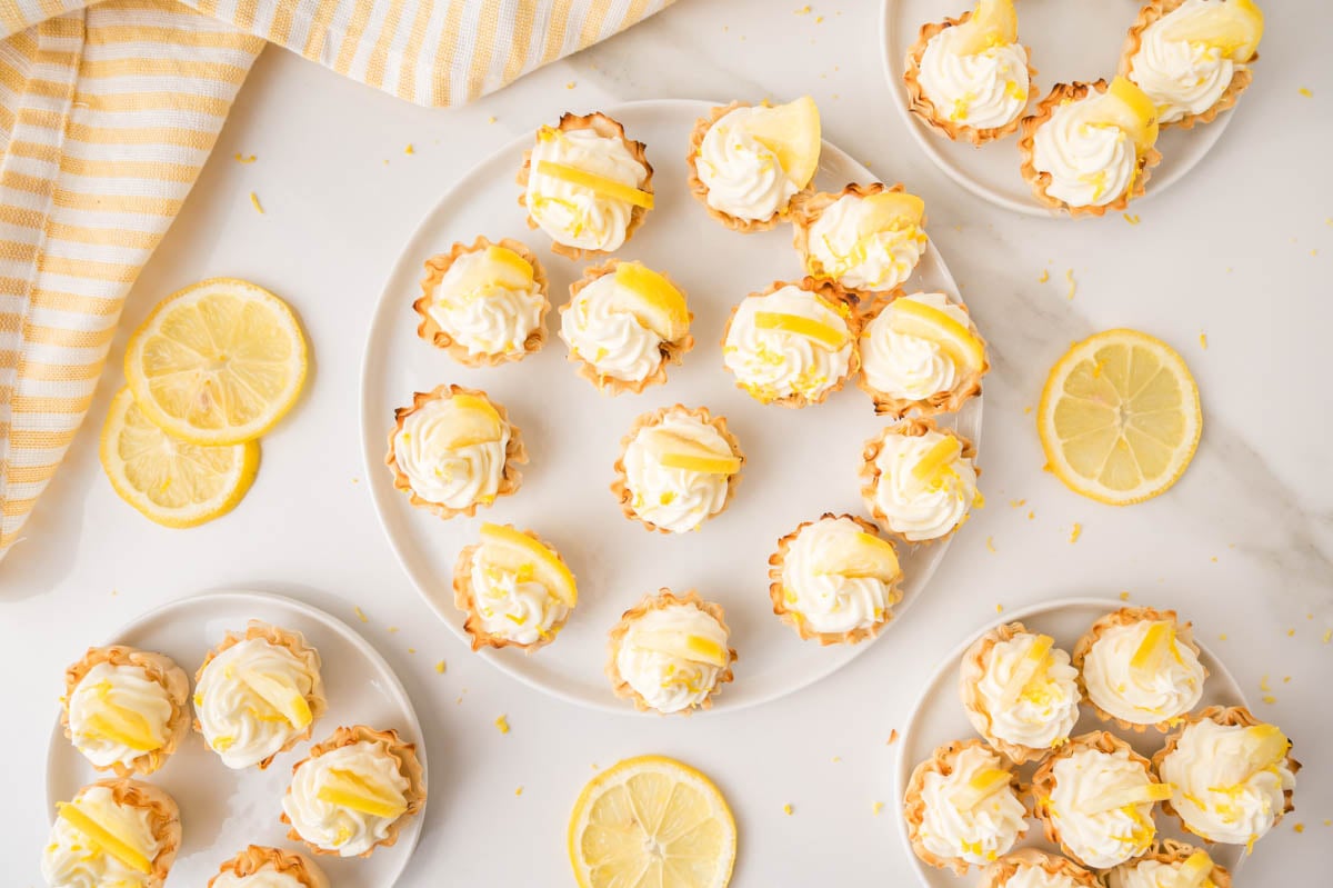 Mini lemon cheesecake bites are arranged on white plates, surrounded by lemon slices and a yellow striped cloth on a white surface.