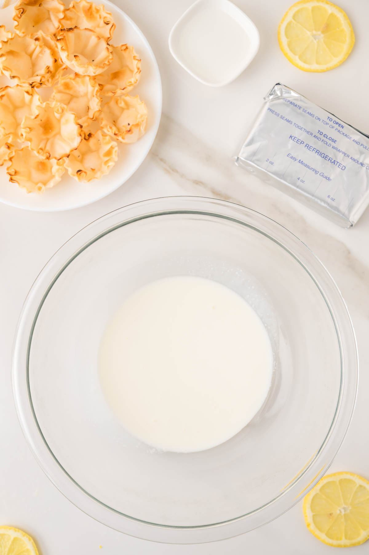 A glass bowl of whipping cream, plate of pastry shells, cream cheese, lemon slices, and a small dish of sugar on a white countertop.