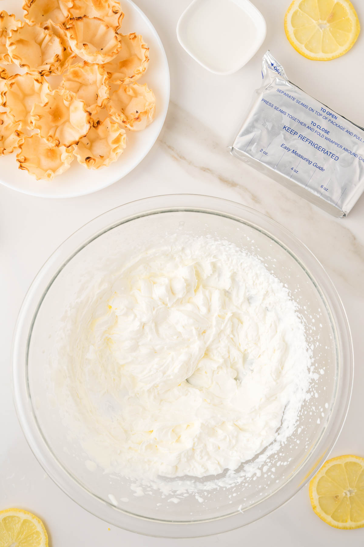 A glass bowl with whipped cream, a plate of empty tart shells, cream cheese packet, lemon slices, and a small cup of milk on a white surface.