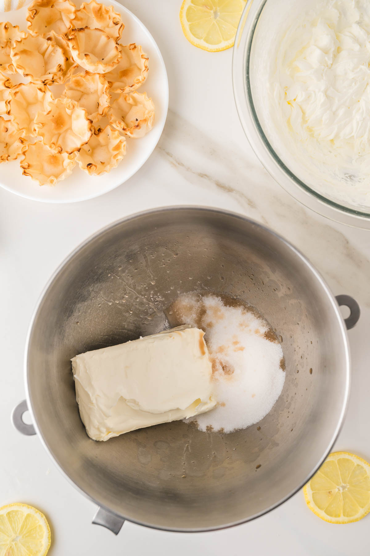 A metal mixing bowl with cream cheese, sugar, and vanilla, surrounded by a plate of pastry shells, a bowl of whipped mixture, and lemon slices on a marble surface.