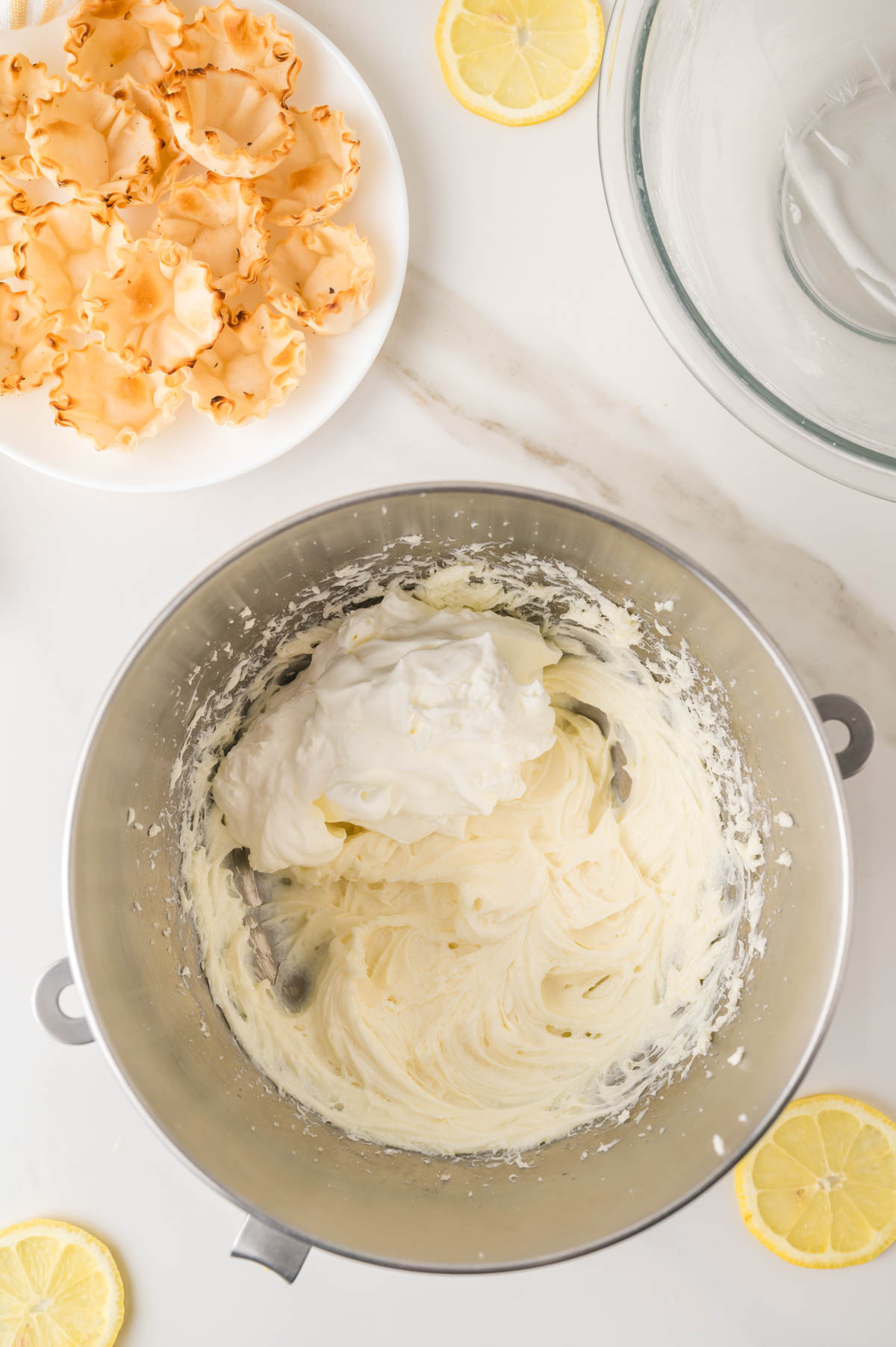 A mixing bowl with a cream cheese mixture and whipped cream, surrounded by empty tart shells on a plate and lemon slices on a white surface.