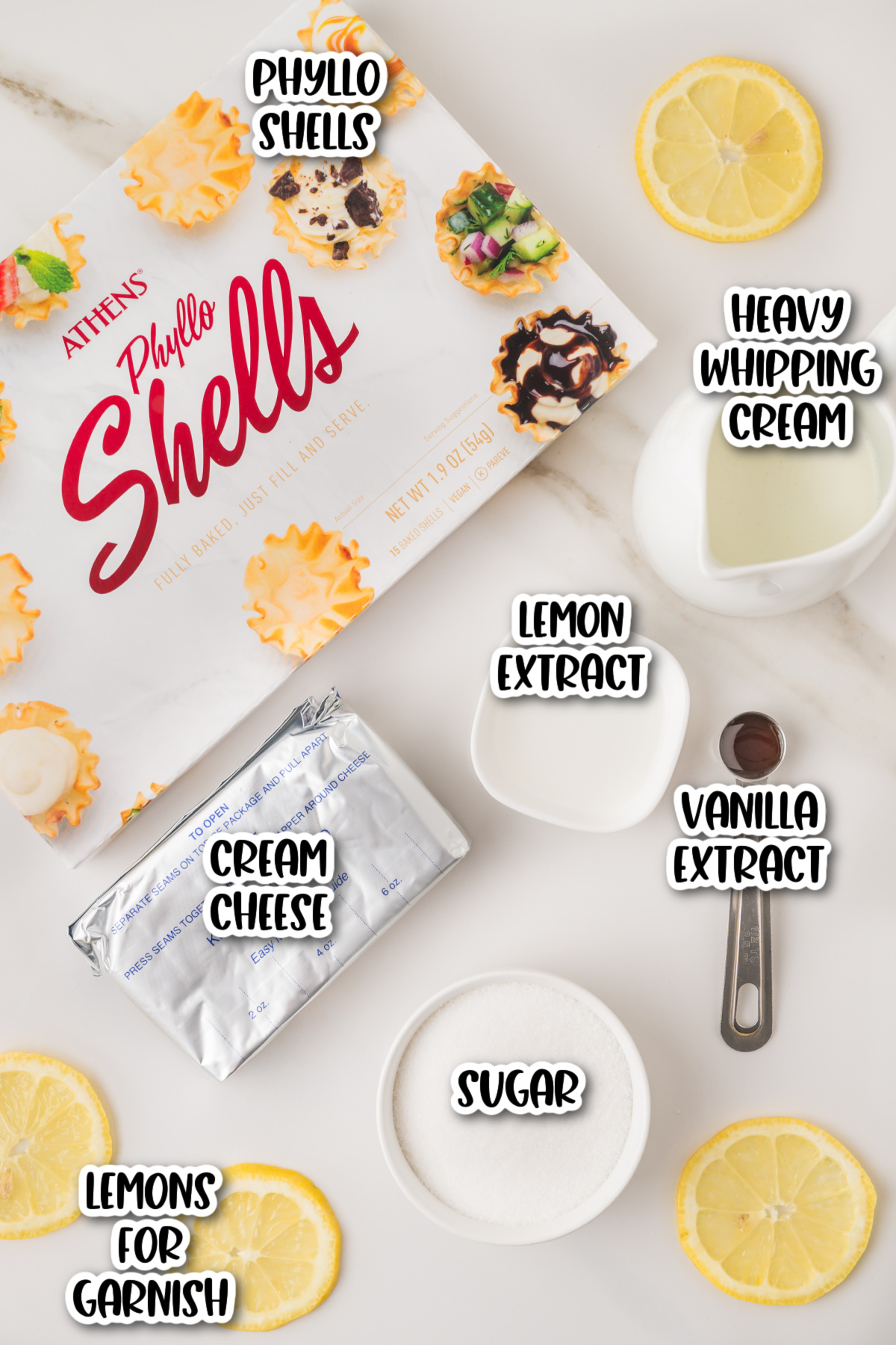 Phyllo shells, cream cheese, sugar, lemon extract, vanilla extract, heavy whipping cream, and lemon slices arranged on a white surface.
