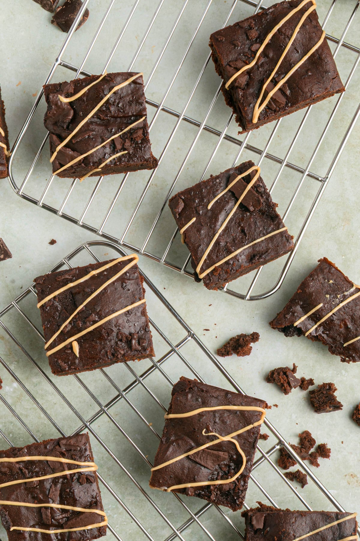 Several chocolate brownies, some drizzled with a light-colored icing, are cooling on wire racks atop a light-colored surface. Crumbs are scattered around.