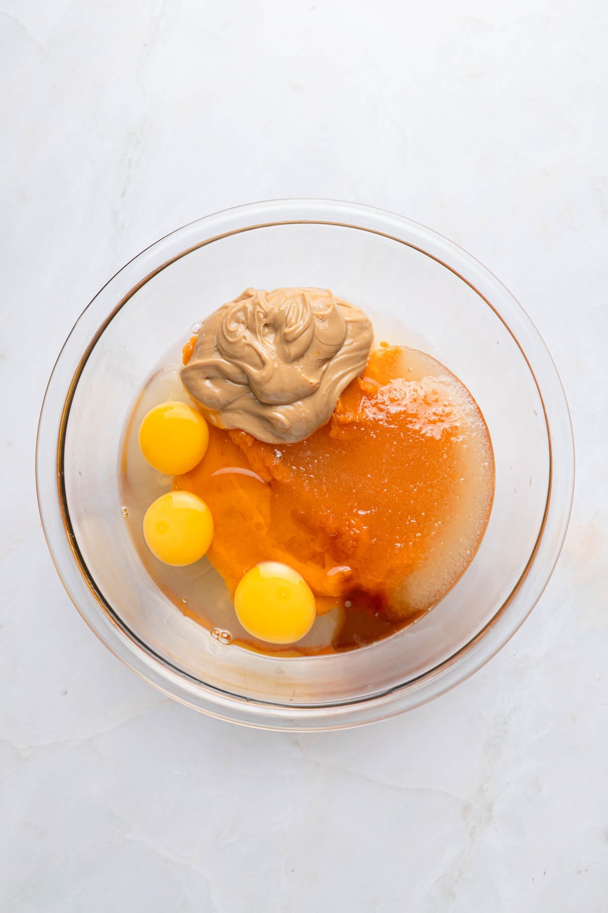 A glass mixing bowl containing three raw eggs, peanut butter, sugar, and pumpkin puree on a white surface.