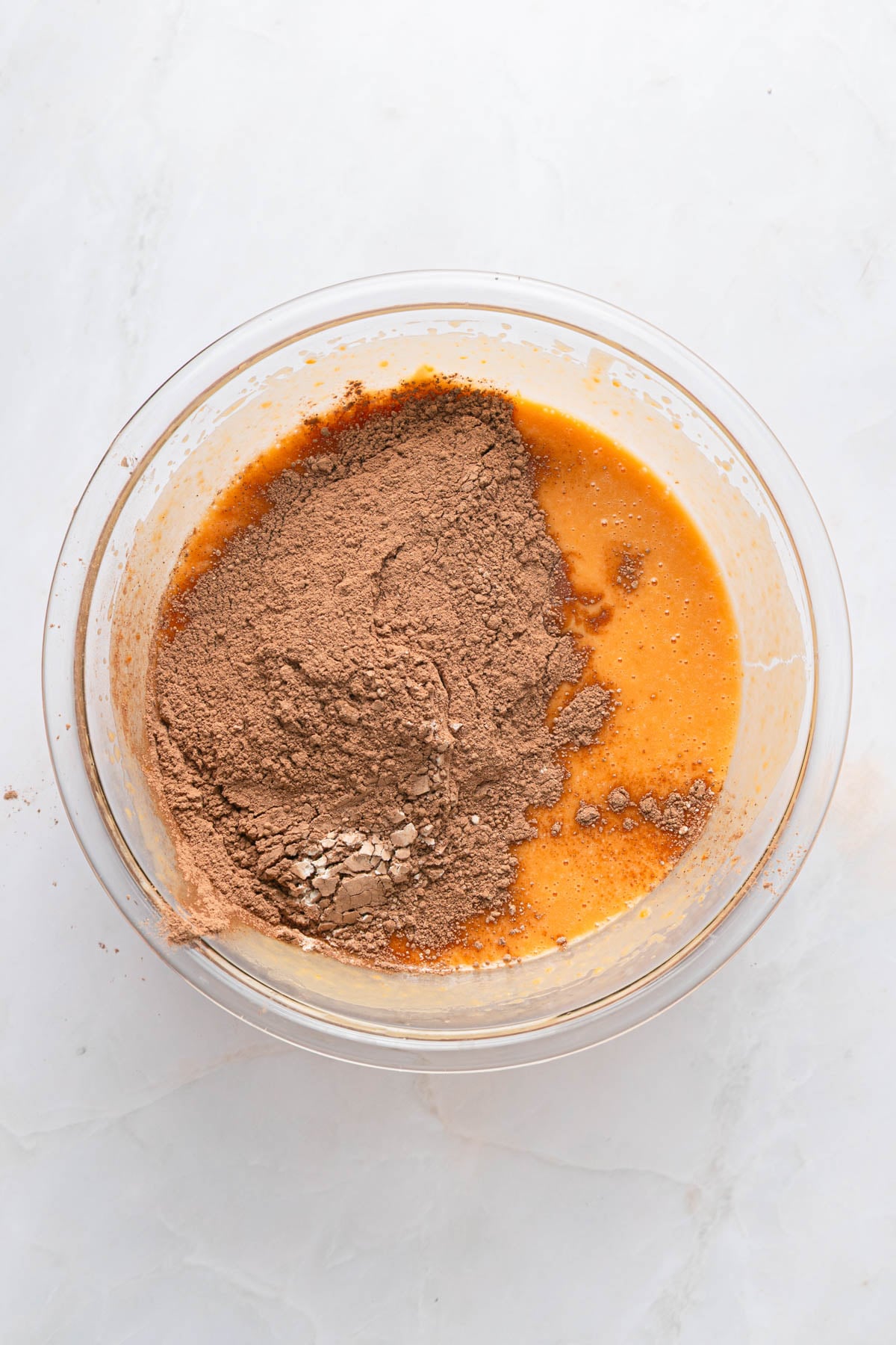 A glass bowl containing a mixture of wet ingredients and cocoa powder on a light surface, ready to be mixed.