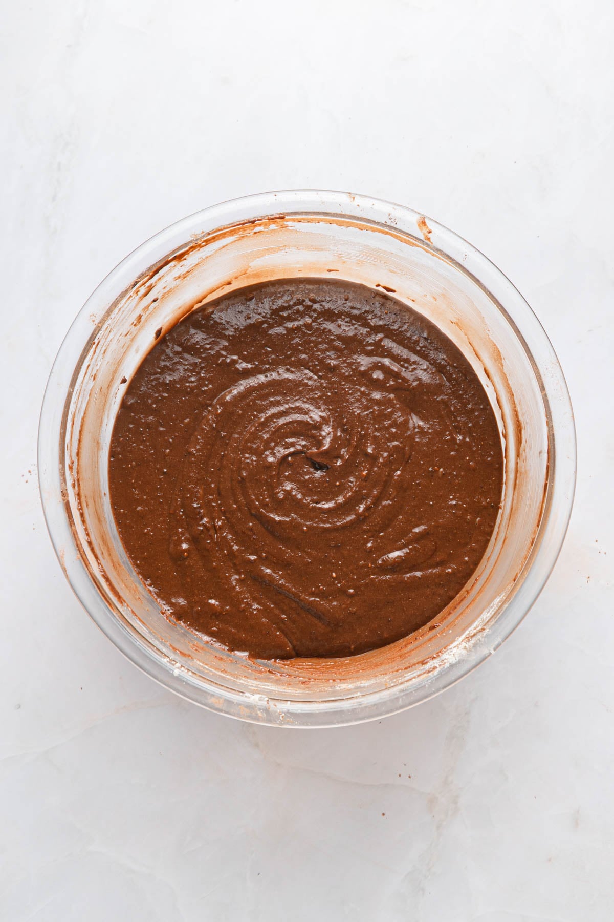 A glass bowl filled with chocolate cake batter sits on a white marble surface.