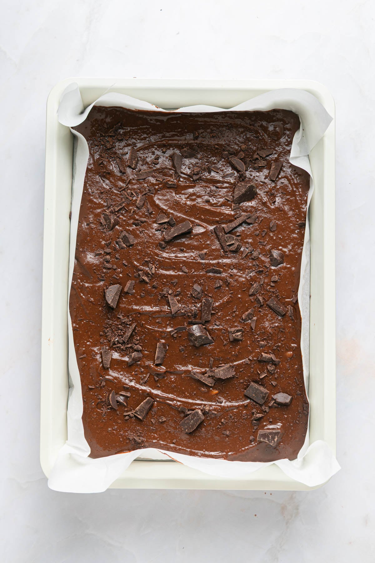 A parchment-lined baking pan filled with chocolate brownie batter, topped with chunks of chocolate, on a white surface.