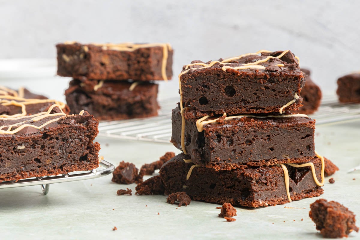 Stacked chocolate brownies with a drizzle of caramel on top, some placed on a cooling rack and others on a light surface with crumbs scattered around.
