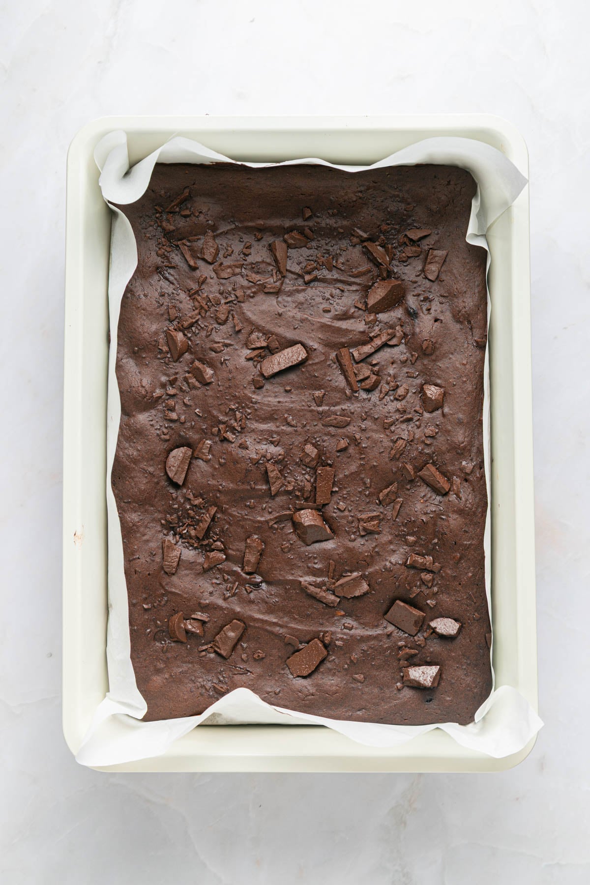 A rectangular baking pan lined with parchment paper holds unbaked chocolate brownie batter topped with chocolate chunks.