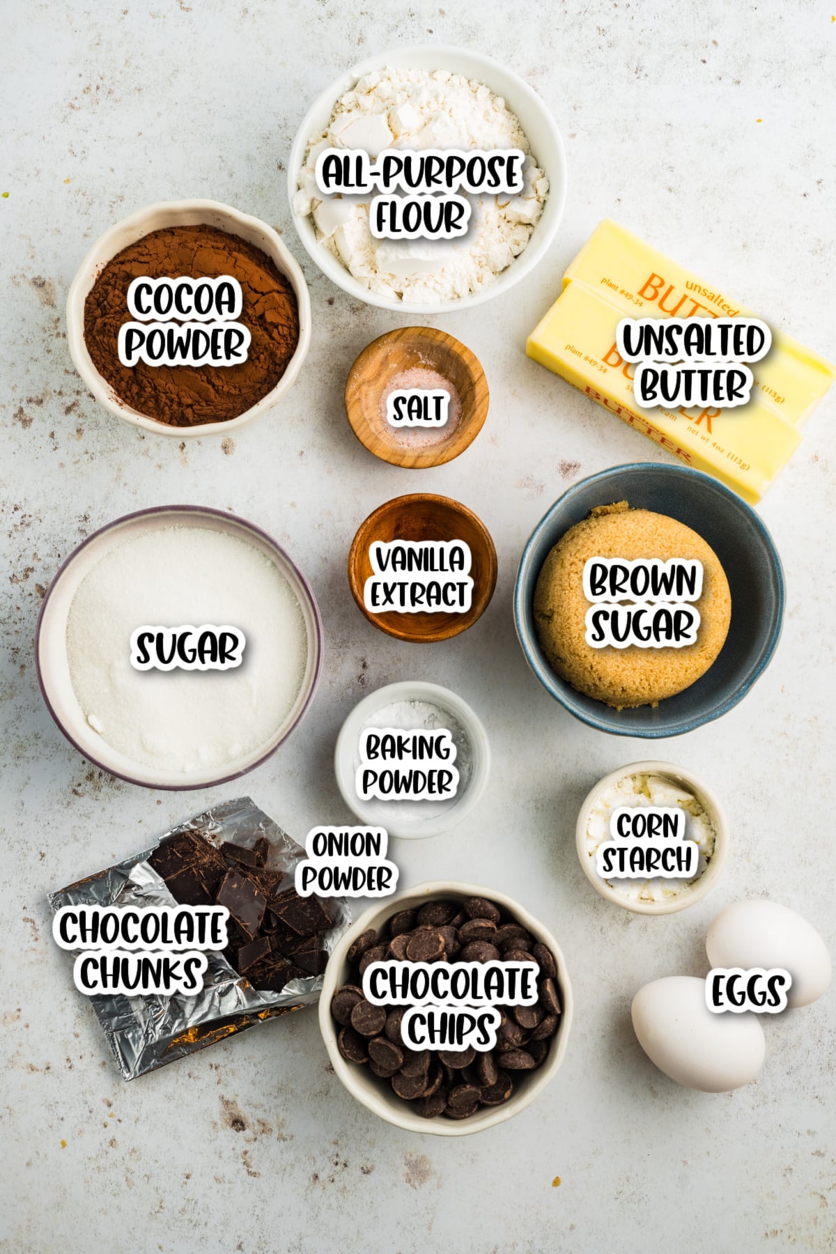 Top-down view of labeled baking ingredients including flour, butter, sugars, cocoa powder, chocolate, eggs, vanilla extract, baking powder, corn starch, onion powder, and salt.