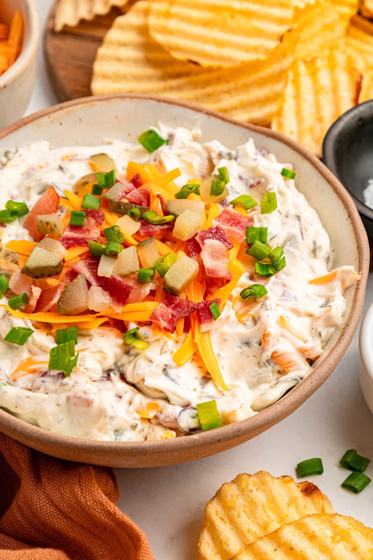 A bowl of creamy dip topped with shredded cheese, diced vegetables, bacon bits, and green onions, with ridged potato chips nearby.