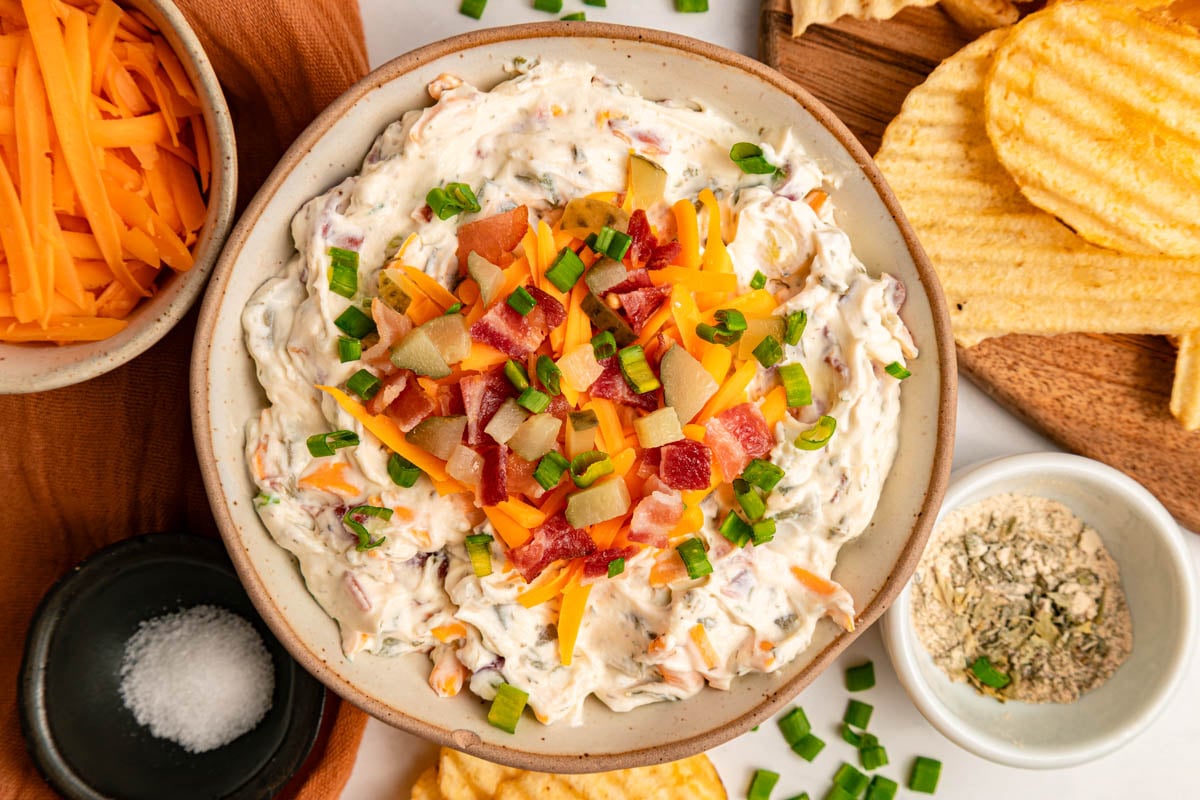 Bowl of loaded baked potato dip topped with cheese, bacon, and green onions, surrounded by potato chips, shredded cheese, and seasoning.