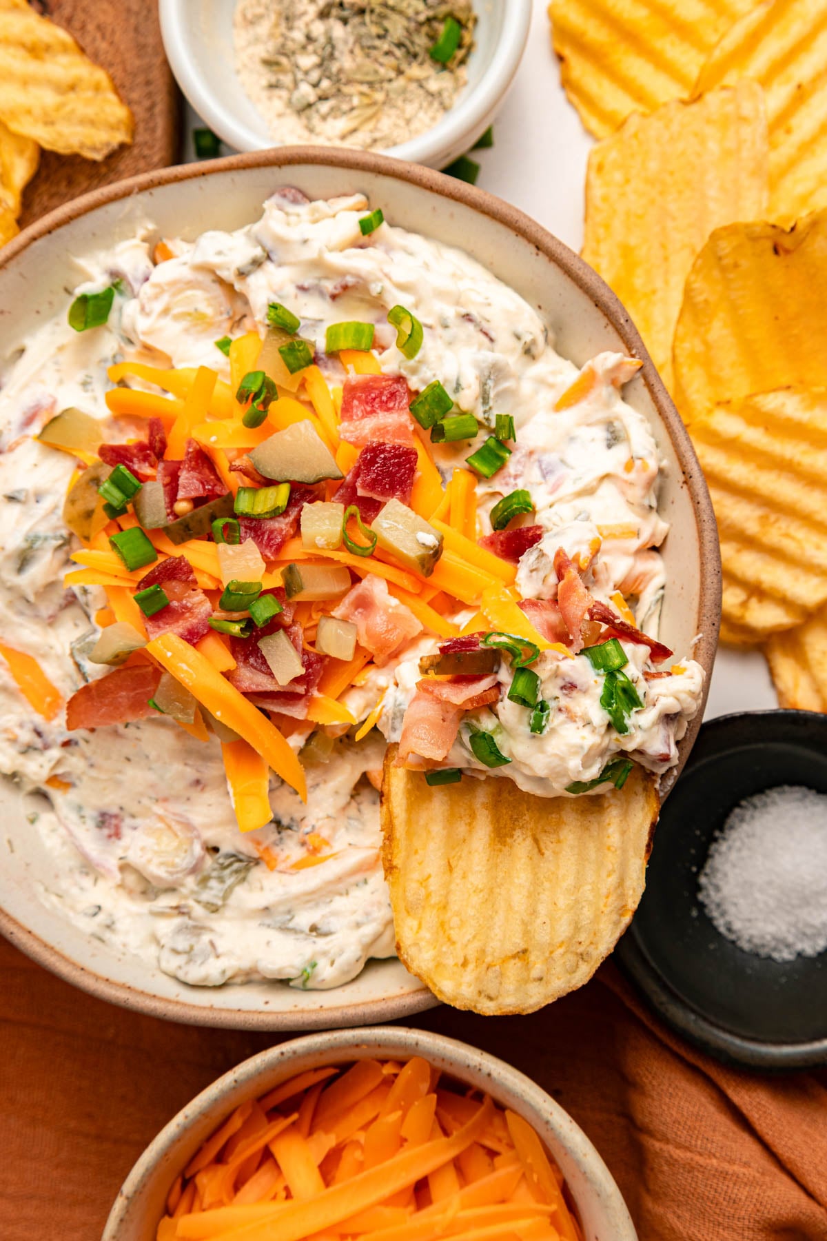 A bowl of creamy dip topped with shredded cheese, bacon, and green onions, surrounded by ridged potato chips and small bowls of seasoning and shredded cheese.